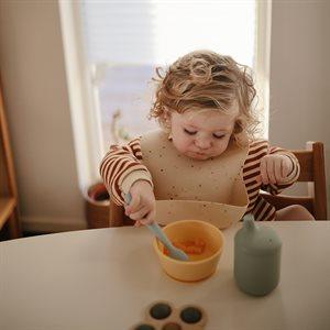 Dziecko, siedzące w krzesełku do karmienia spożywa posiłek niebnieską łyżeczką, marki Mushie.